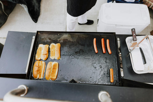 Overhead view of hot dogs and buns grilling at an outdoor BBQ setting.