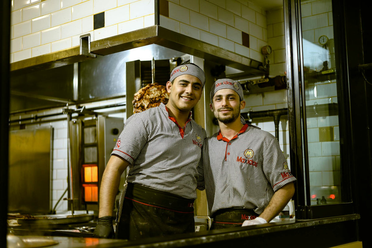 Two chefs in uniforms smiling in an industrial kitchen environment, showcasing teamwork and culinary expertise.