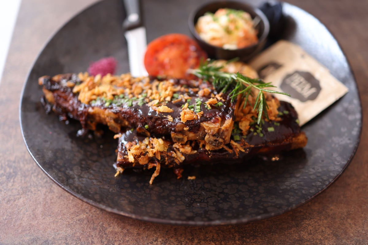 Close-up of a gourmet ribs dish topped with crispy onions and fresh herbs, served on a dark plate.