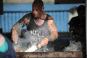 A tattooed chef grills food outdoors, surrounded by smoke and natural light.