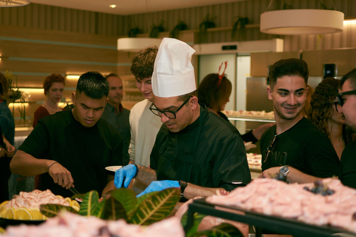 A chef in glasses serves food to guests at a lively indoor buffet event.