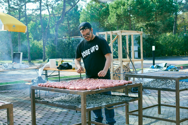A man grills sausages at an outdoor event with trees in the background.