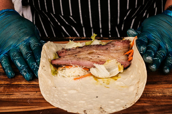 Chef uses protective gloves to prepare a beef wrap with fresh ingredients on a wooden surface.