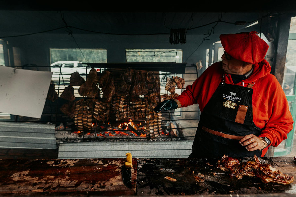 Man preparing traditional Argentine asado with grilled meat on open fire outdoors.