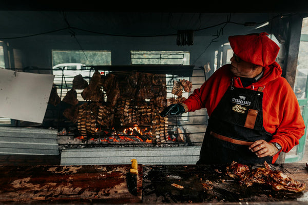 Man preparing traditional Argentine asado with grilled meat on open fire outdoors.