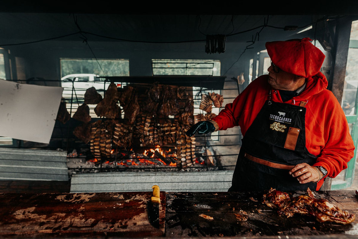 Man preparing traditional Argentine asado with grilled meat on open fire outdoors.