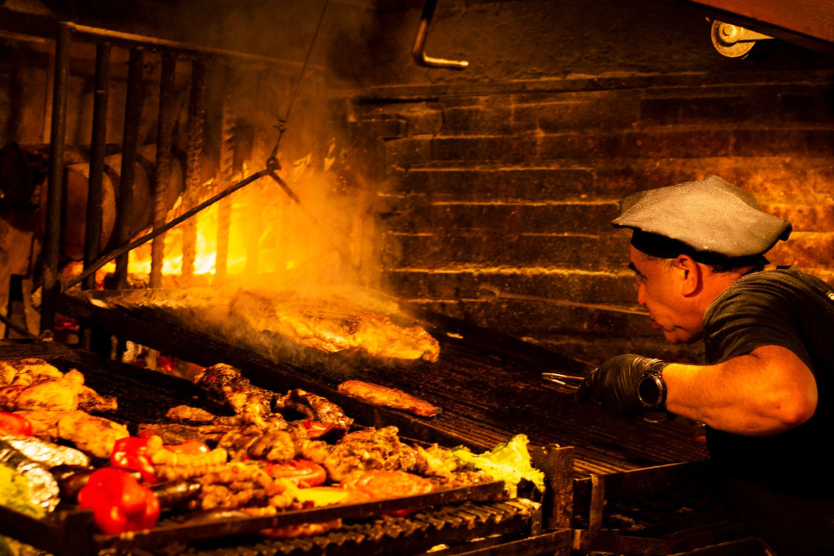 Chef prepares a traditional asado barbecue with sizzling meats over an open fire grill.