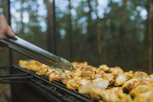 A close-up of meat being grilled outdoors in a forest setting with tongs.