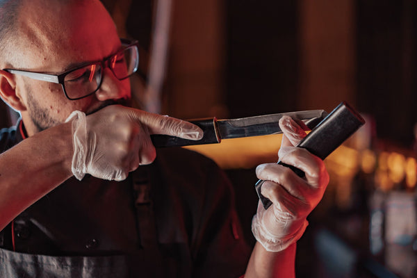 A chef sharpening a knife with precision in a warm-lit kitchen, wearing latex gloves and glasses.