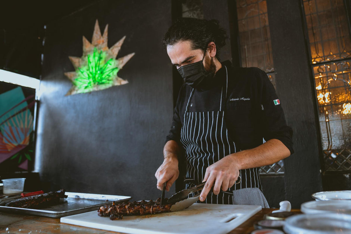 A chef wearing a mask slices grilled steak on a chopping board indoors.